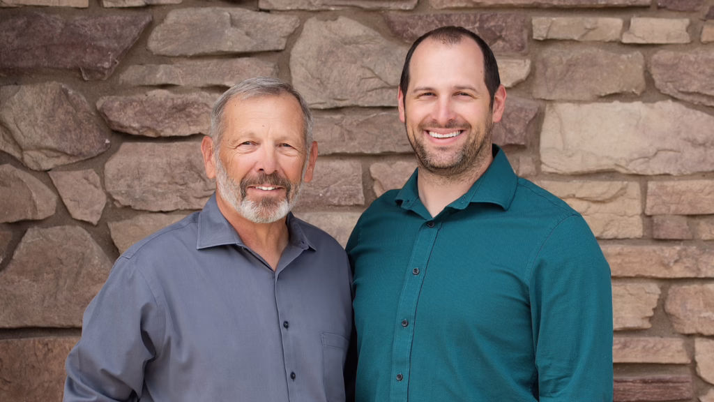 Two men standing side by side and smiling in front of a stone wall backdrop.